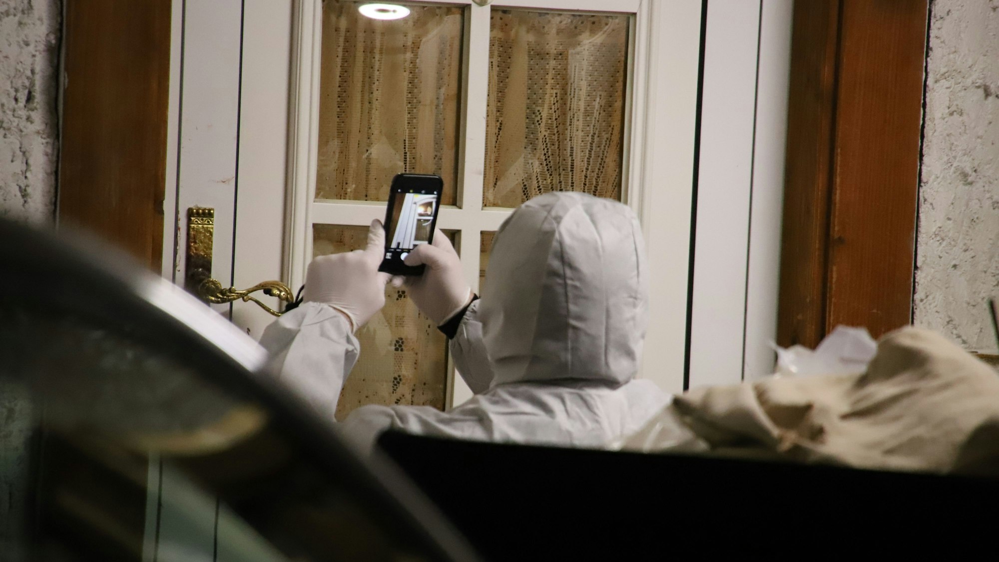 Ein Polizist der Spurensicherung im weißen Overall mit Kapuze fotografiert mit dem Handy eine Haustür. Es ist Nacht.