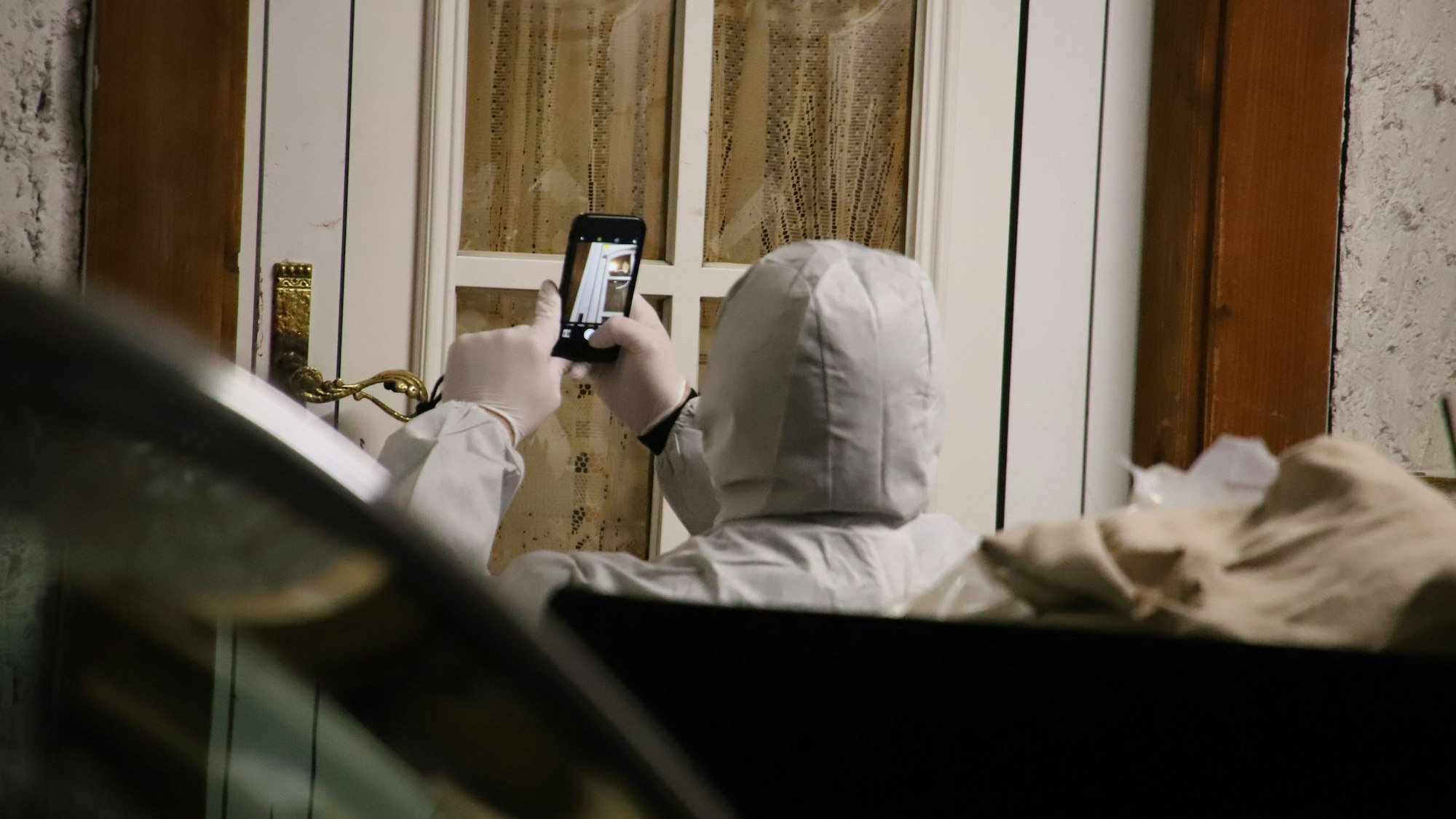 Eine Person in einer weißen Schutzkleidung macht Fotos von einer Eingangstür mit dem Handy.