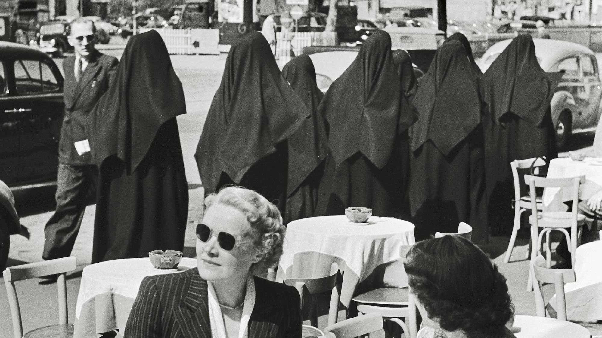 Mehrere Nonnen gehen an einer Straße, im Vordergrund sitzen zwei Frauen.