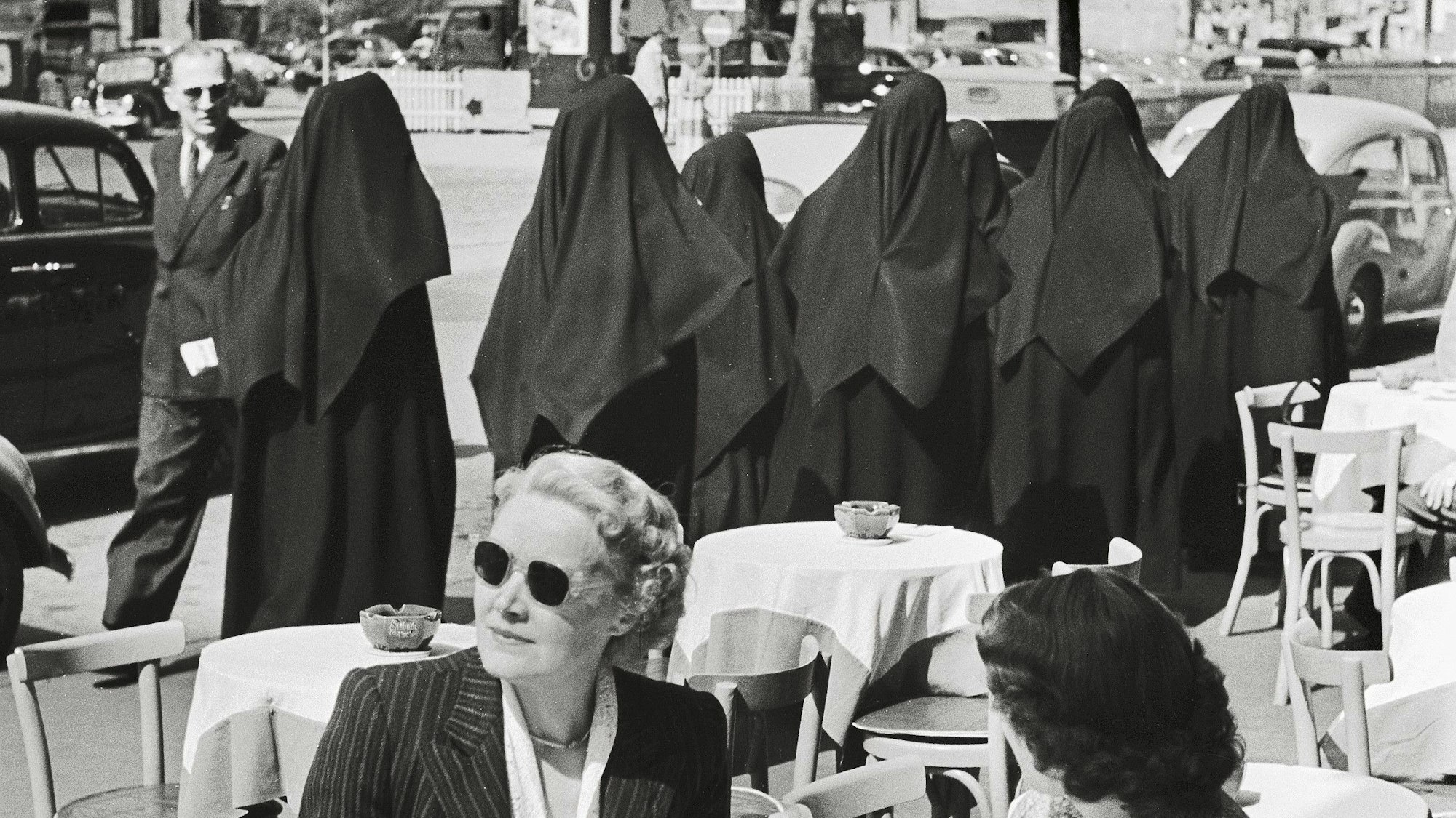 Mehrere Nonnen gehen an einer Straße, im Vordergrund sitzen zwei Frauen.
