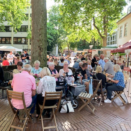 Das Foto zeigt den Schlemmermarkt in Frechen. Besucher sitzen an Tischen und genießen das kulinarische Angebot.