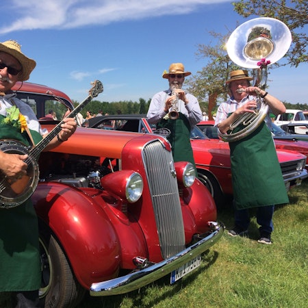Es sind drei Männer zu sehen, die vor einem Oldtimer Musik spielen.
