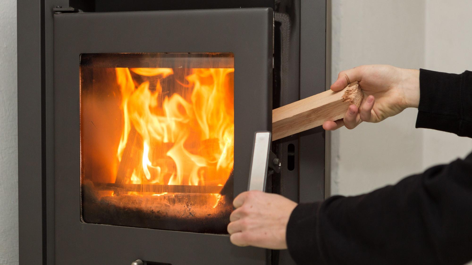 Ein Mann legt in seiner Wohnung ein Stück Holz in einen Kaminofen, in dem ein Feuer brennt.