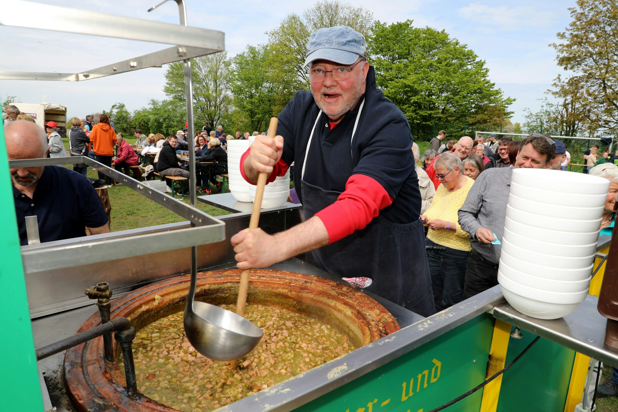 Ein Mann mit Kappe rührt mit einem langen Holzlöffel in einem großen Topf Erbsensuppe, während Menschen im Hintergrund Schlange stehen.