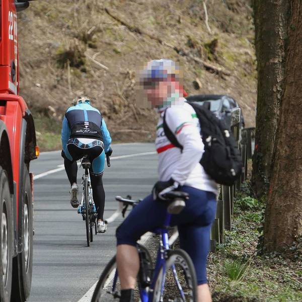 Ein Rennradfahrer mit Rucksack wartet am Fahrbahnrand, bis ein roter Betonmischer vorbeigefahren ist. Vor dem Lkw ist ein weiterer Radfahrer auf der L 144 unterwegs.