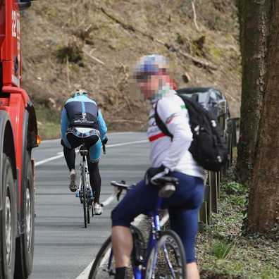 Ein Rennradfahrer mit Rucksack wartet am Fahrbahnrand, bis ein roter Betonmischer vorbeigefahren ist. Vor dem Lkw ist ein weiterer Radfahrer auf der L 144 unterwegs.