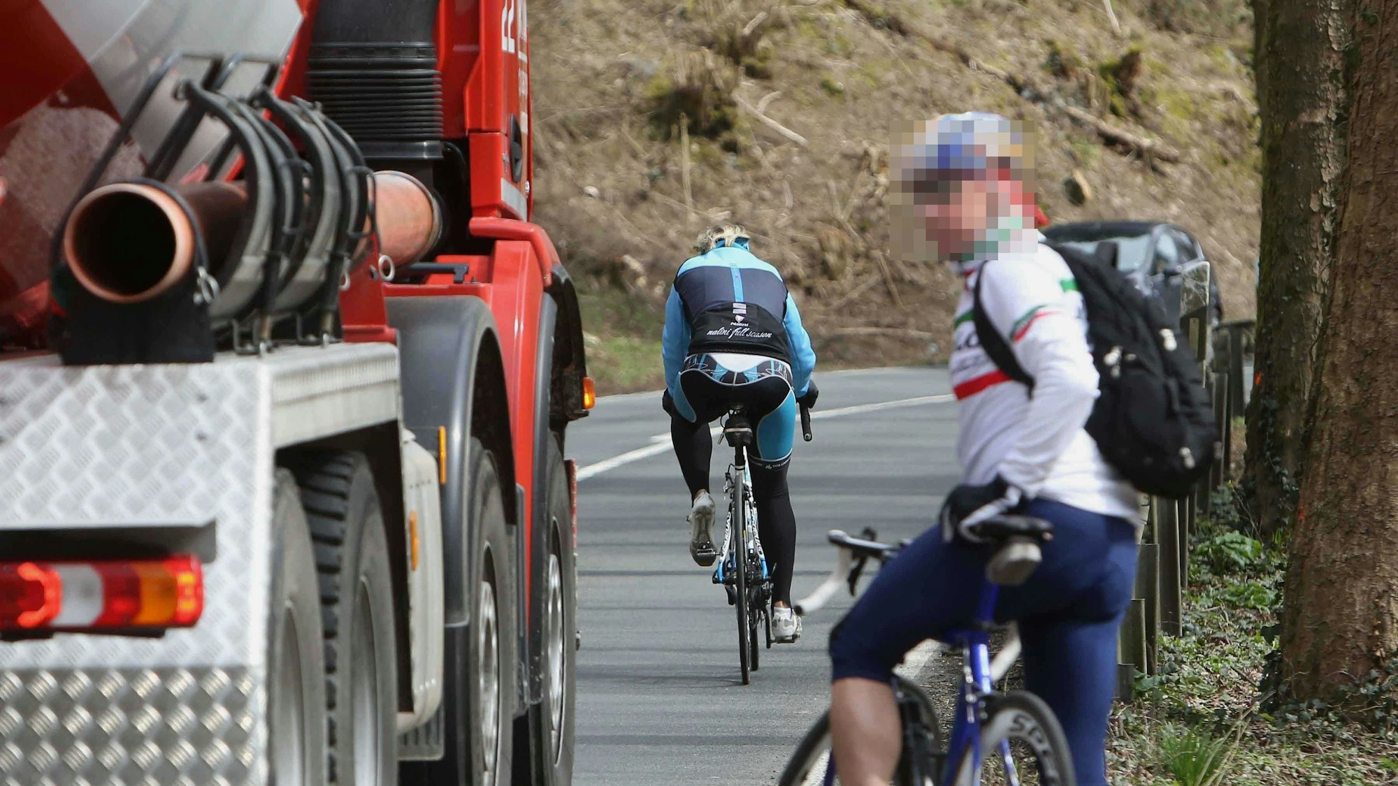 Ein Rennradfahrer mit Rucksack wartet am Fahrbahnrand, bis ein roter Betonmischer vorbeigefahren ist. Vor dem Lkw ist ein weiterer Radfahrer auf der L 144 unterwegs.
