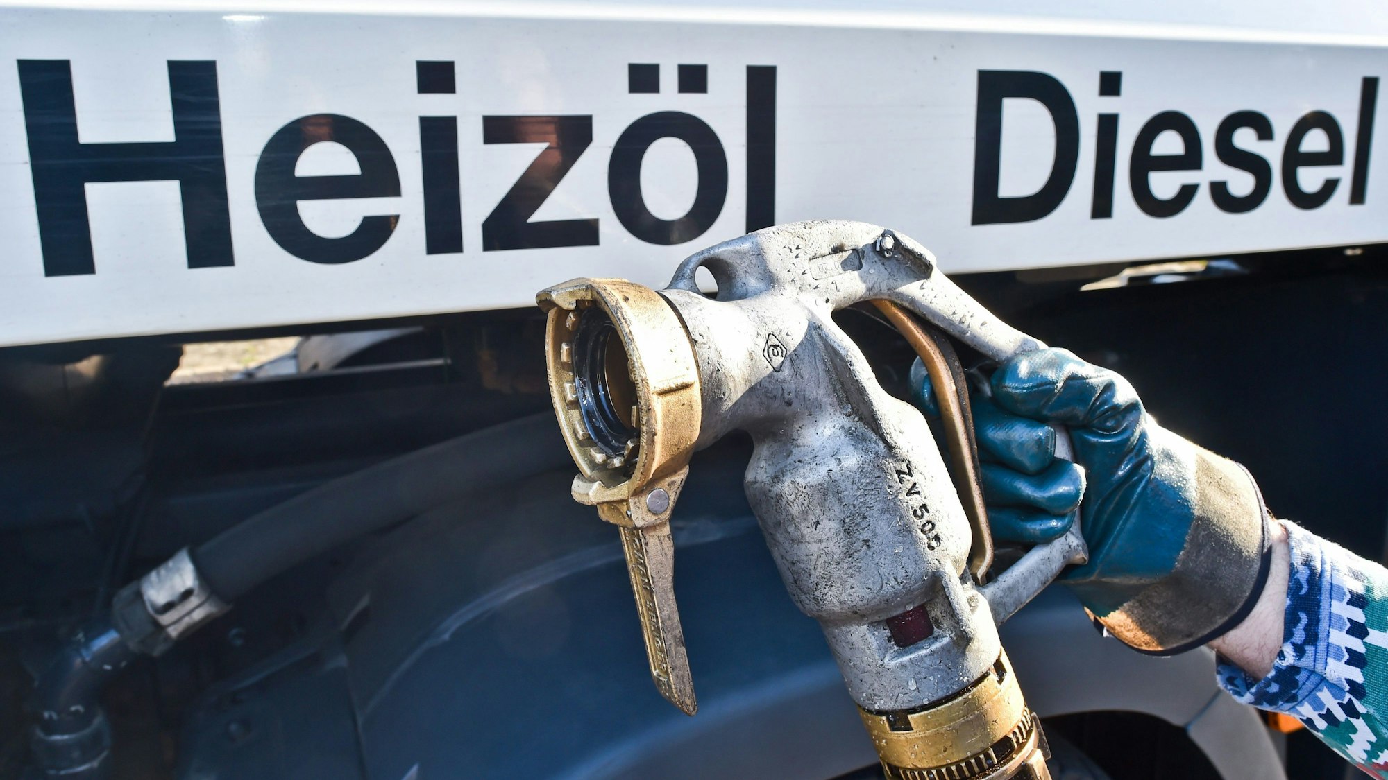 Eine Hand mit Handschuh hält einen Zapfhahn für Heizöl vor dem Ausschnitt eines Heizöllieferanten.