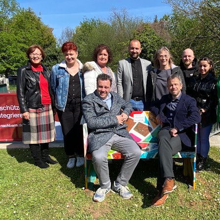 Es sind mehrere Menschen zu sehen, die um die Bank herumstehen. Zwei von ihnen sitzen auf ihr drauf.