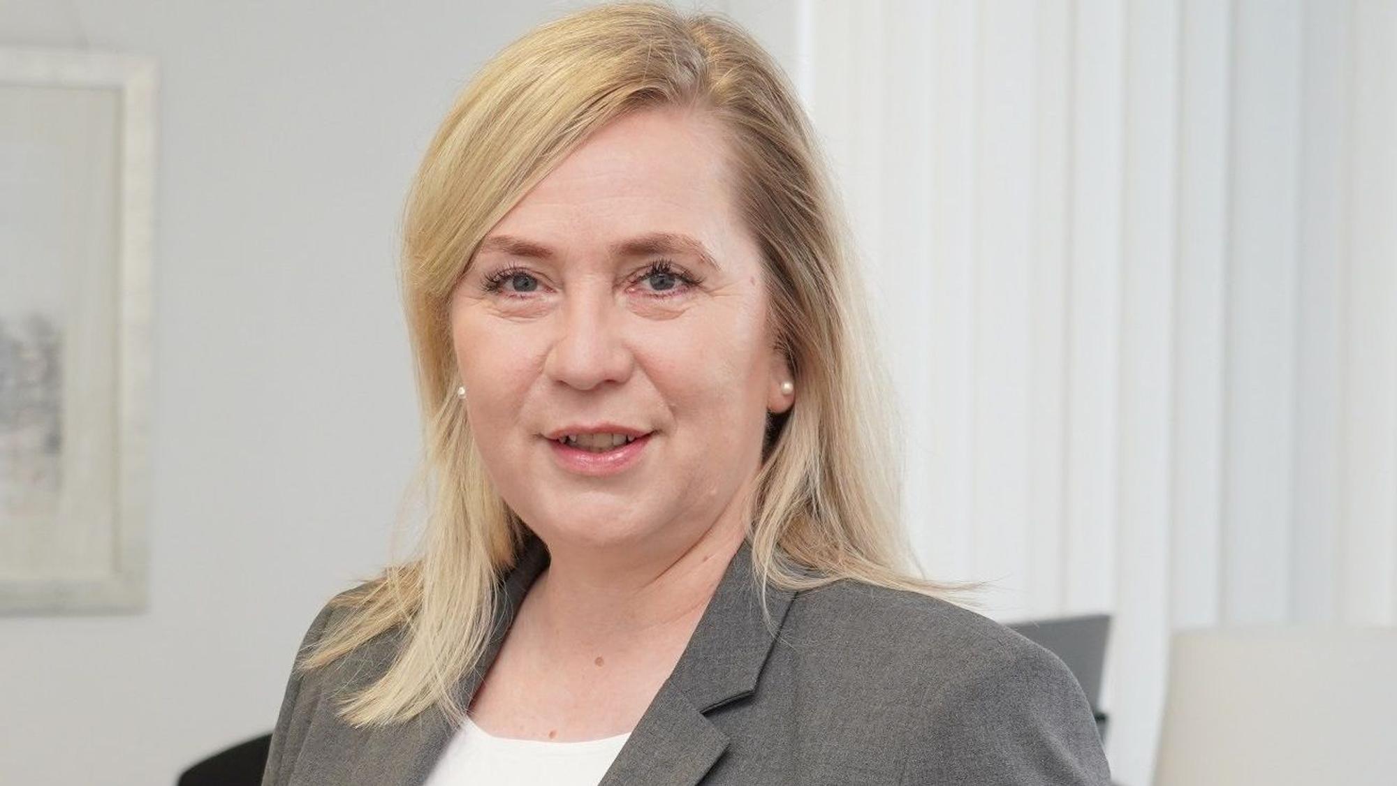 Auf dem Foto ist eine Frau mit blondem Haar zu sehen. Sie trägt einen grauen Blazer.