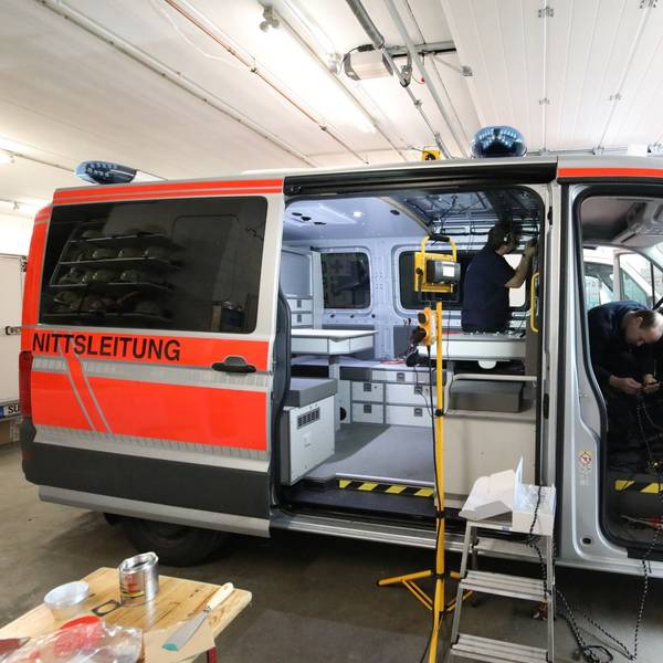 Ein Transporter steht mit offener Schiebetür und geöffneter Beifahrertür in einer Halle. Zwei Männer arbeiten in dem Fahrzeug, das durch einen Scheinwerfer ausgeleuchtet wird.