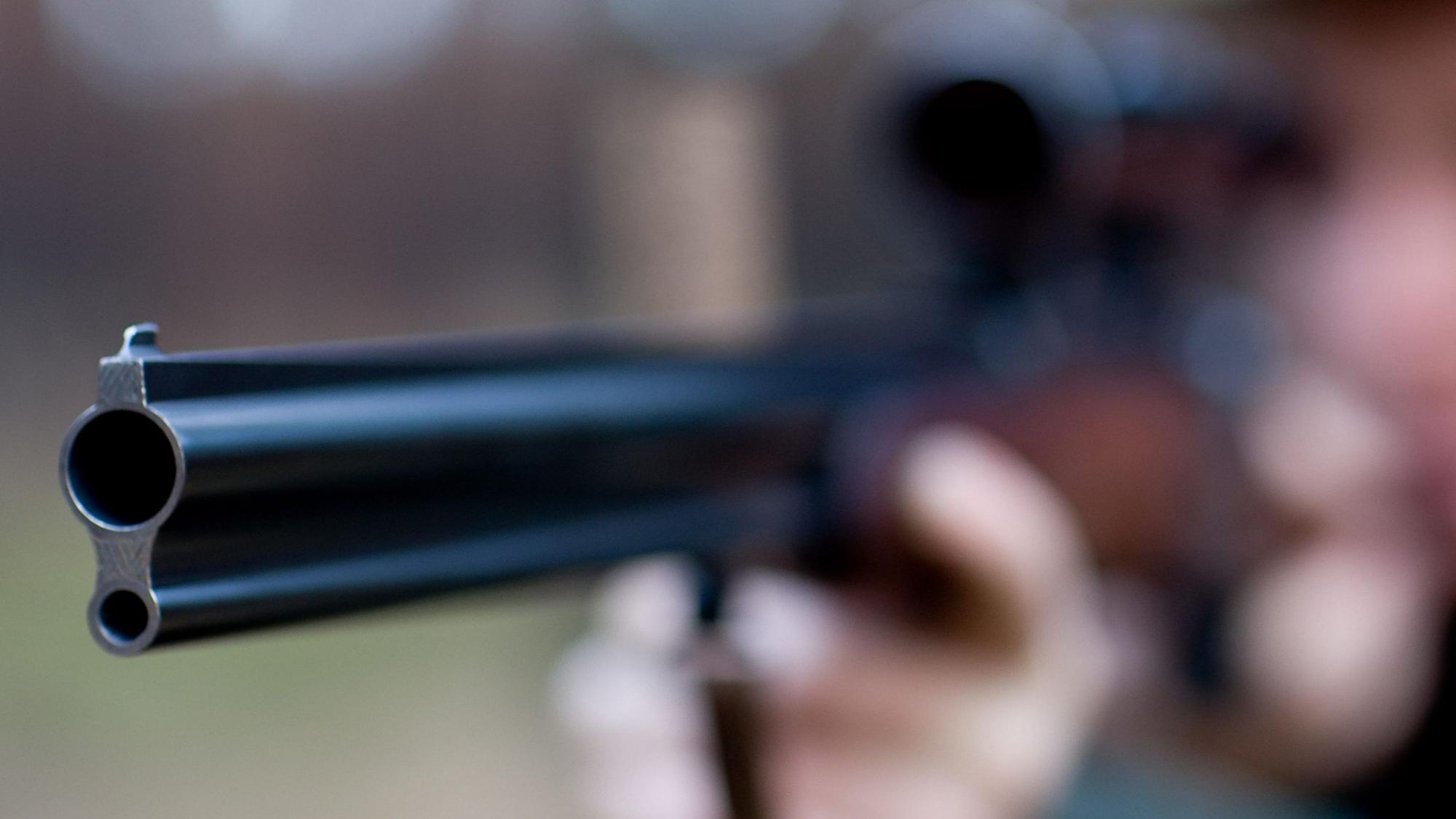 Ein im Hintergrund nur verschwommen zu erkennender Mann zielt durch ein Fernrohr mit einem Gewehr.