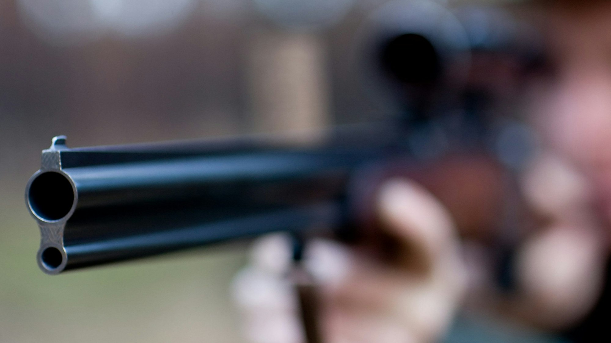 Ein im Hintergrund nur verschwommen zu erkennender Mann zielt durch ein Fernrohr mit einem Gewehr.