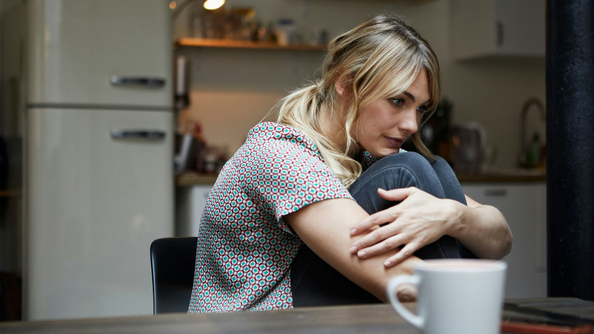 Eine Frau sitzt etwas zusammengekauert alleine an einem Tisch und starrt ins Leere