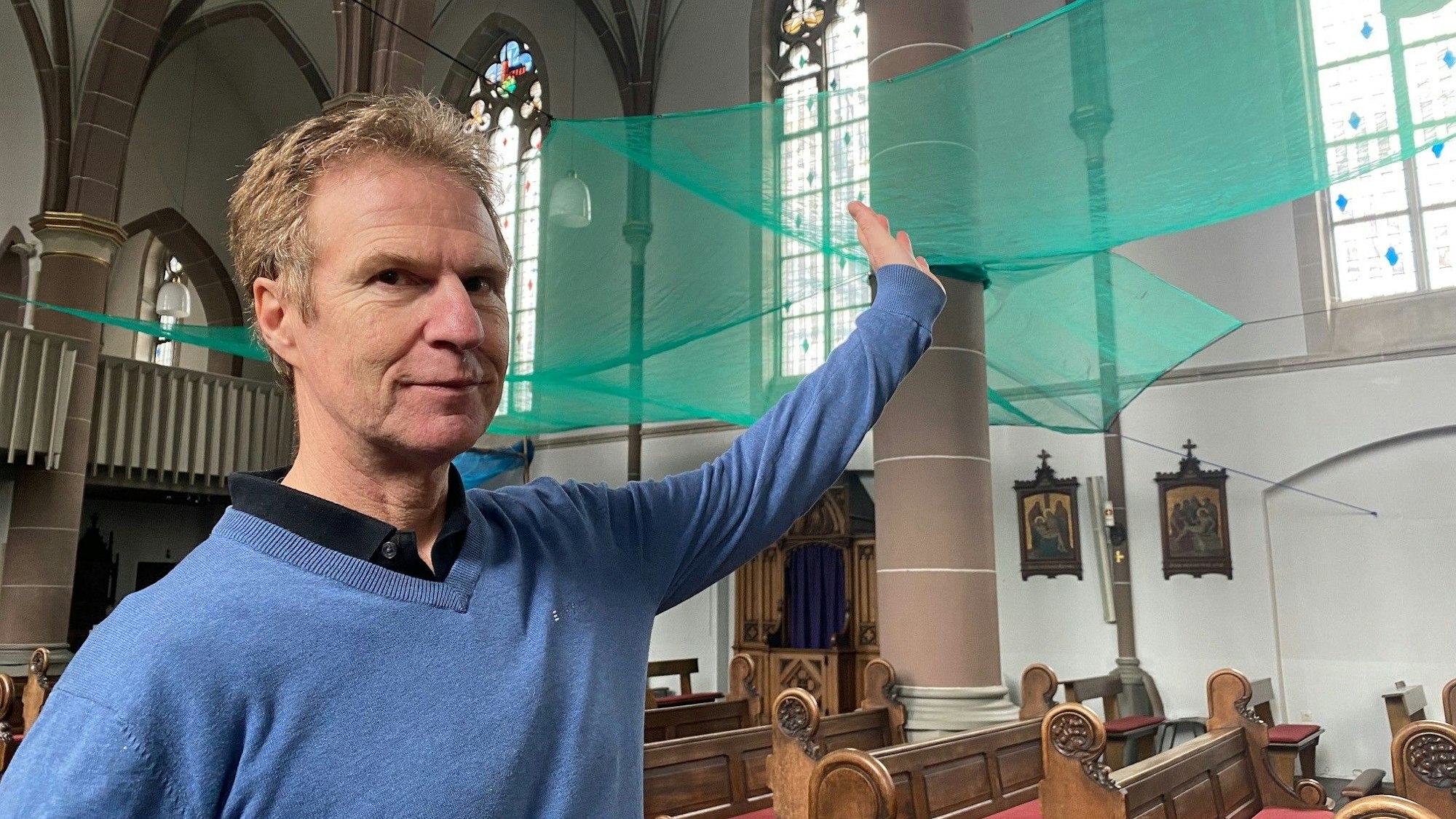 Felix Ross trägt einen blauen Pullover, blickt in die Kamera und zeigt auf das grüne Netz.