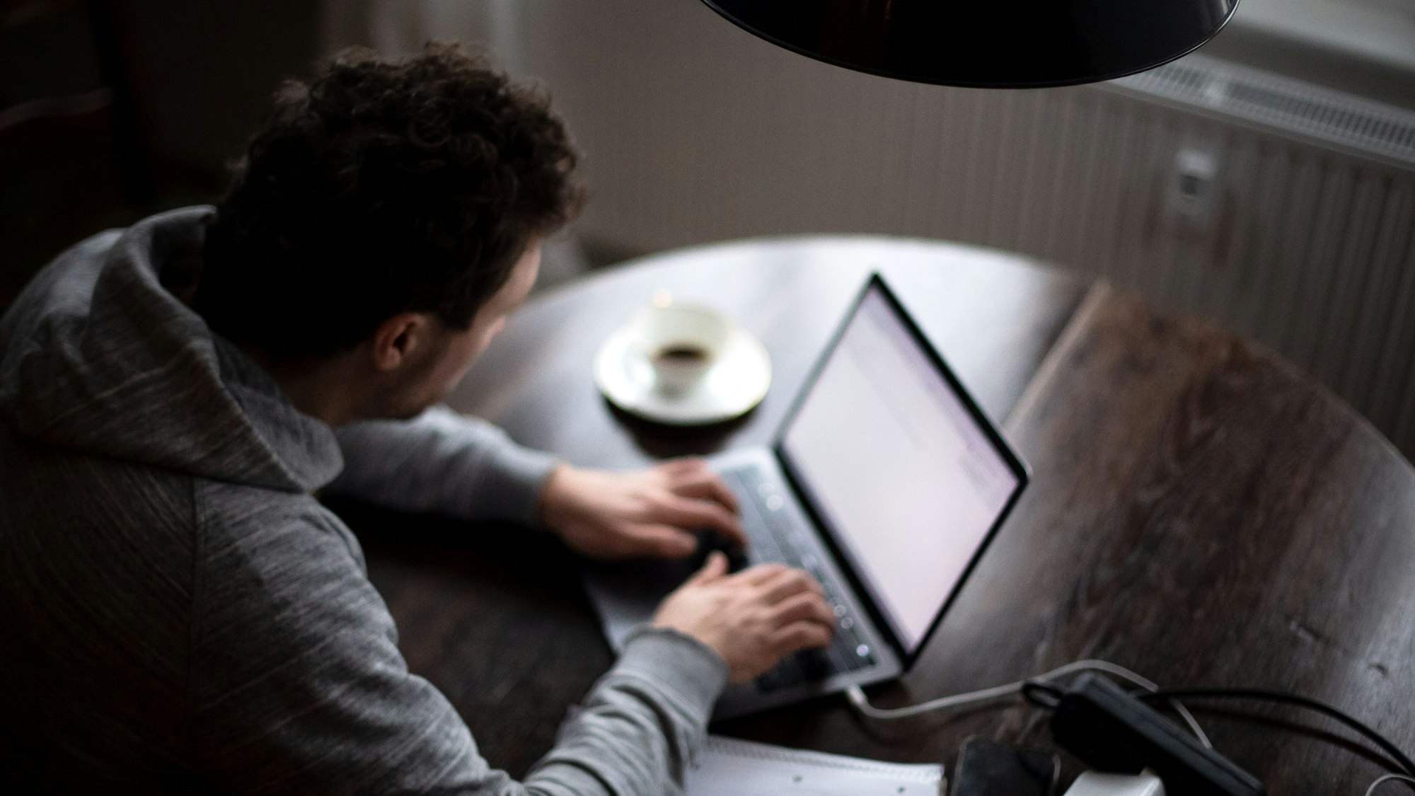 Ein Mann sitzt an einem runden Tisch und tippt auf einem Laptop. Neben ihm steht eine Tasse mit Kaffee.