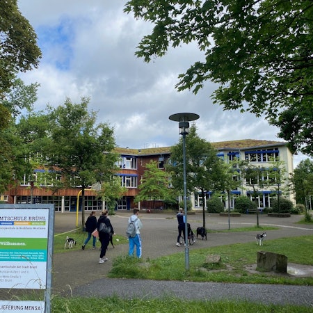 Blick auf den Schulhof der Gesamtschule. Die Gebäude werden zum Teil von grünen Bäumen verdeckt. Zwei Schüler und zwei Spaziergänger mit ihren Hunden laufen vorbei.