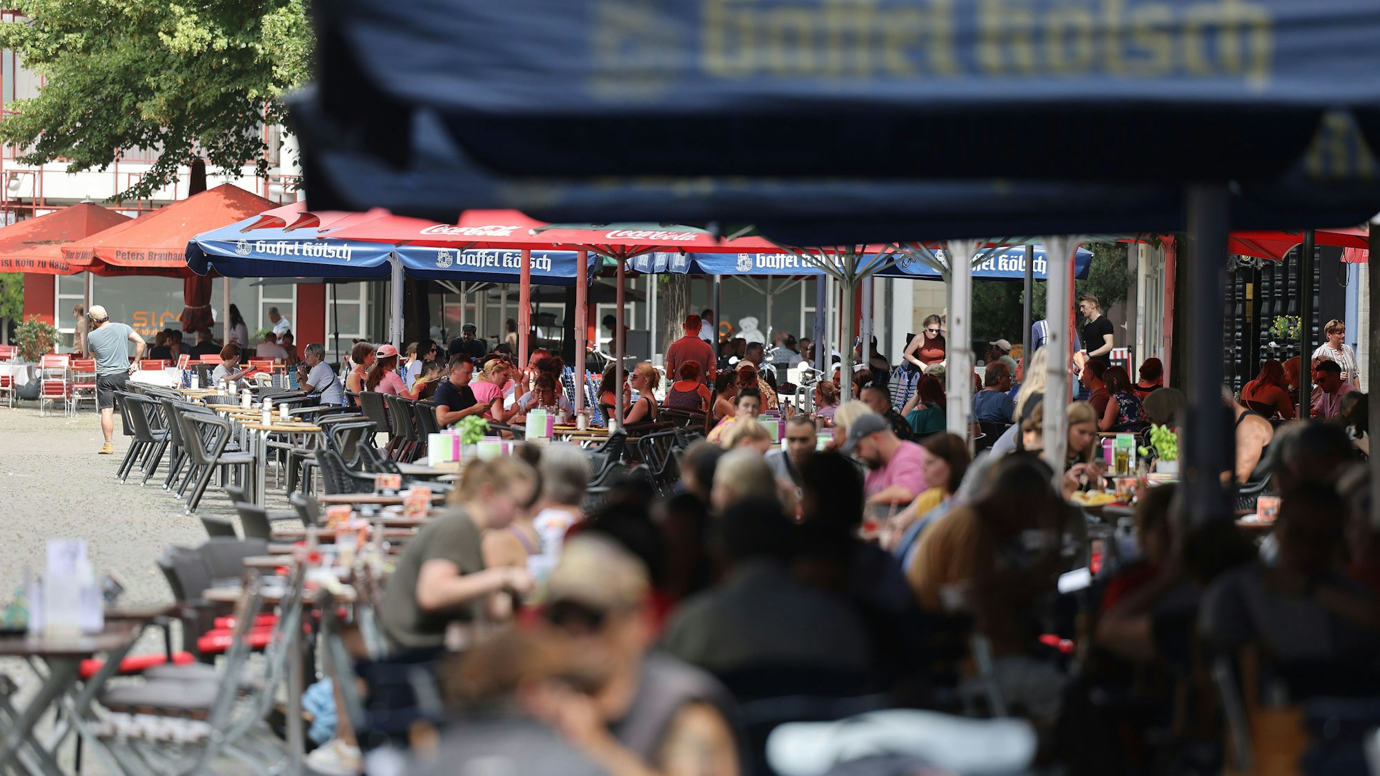 Menschen sitzen in Köln in Lokalen im Außenbereich unter Sonnenschirmen. Die Bierpreise könnten 2023 deutlich steigen, das erwarten Brauerei-Verbände. (Archivbild)