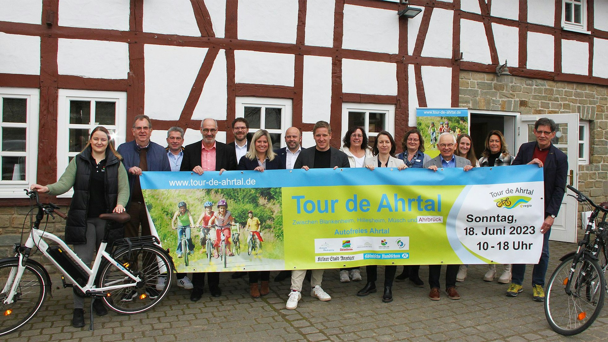 Die Vertreter der beteiligten Kreise und Kommunen halten vor einem Fachwerkgebäude in Antweiler ein Banner, mit dem für die „Tour de Ahrtal“ geworben wird.