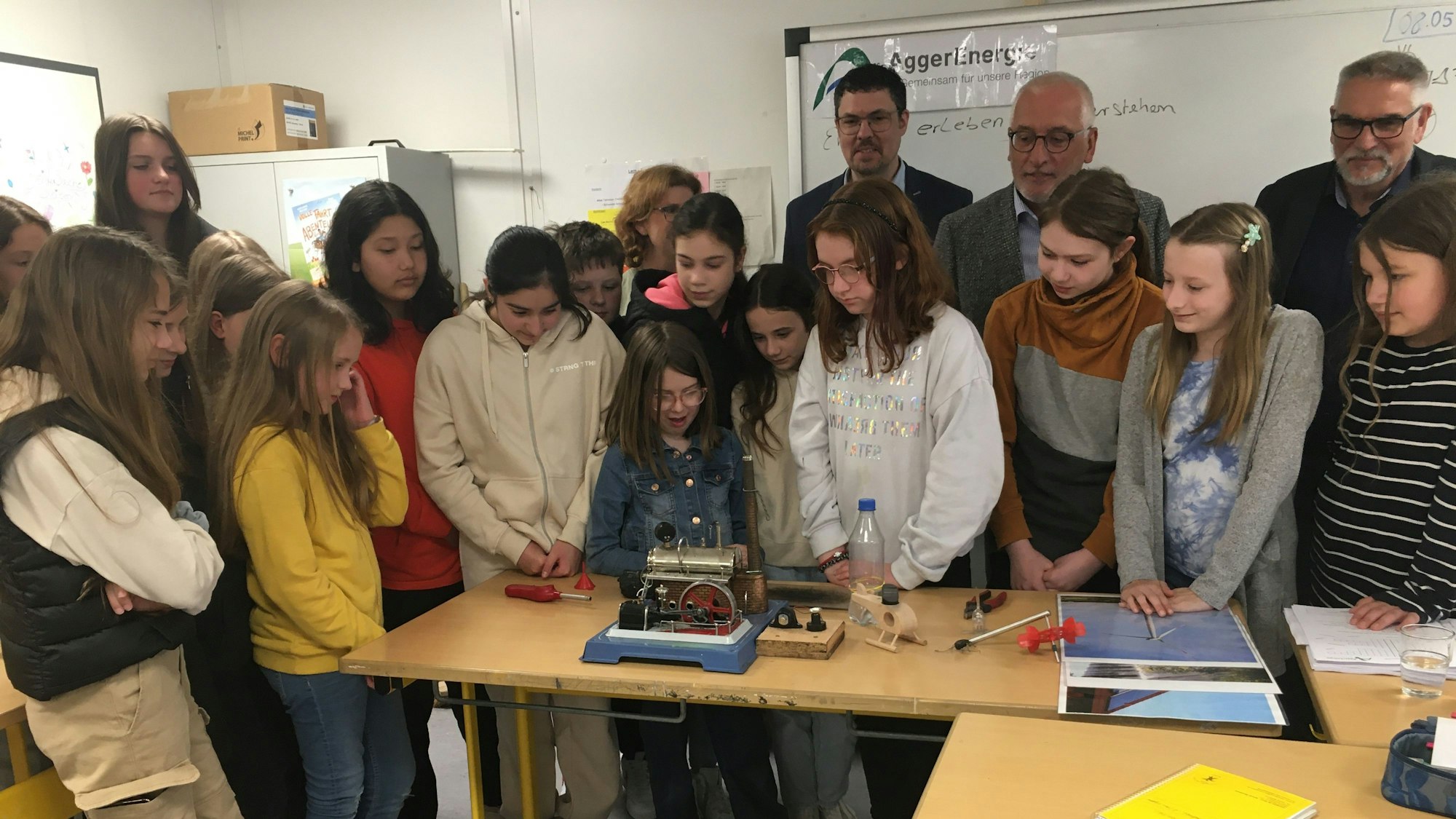 Eine Schulklasse steht als Gruppe neugierug vor einem Experiment, das auf dem Tisch vor ihr aufgebaut ist.