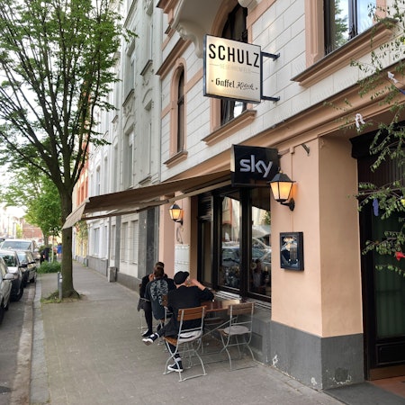 Außenansicht des Lokals Schulz in der Landmannstraße in Neuehrenfeld mit geschmücktem Maibaum und Gästen.
