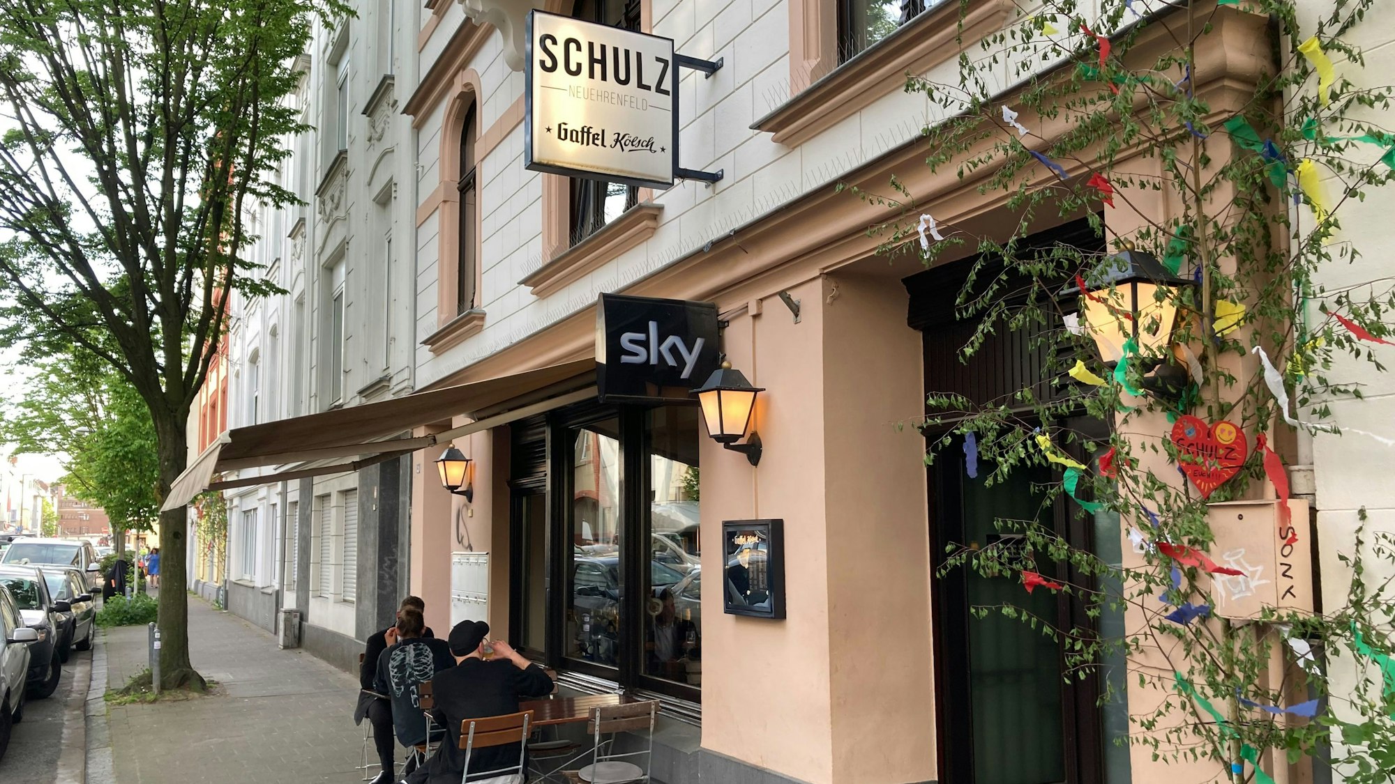 Außenansicht des Lokals Schulz in der Landmannstraße in Neuehrenfeld mit geschmücktem Maibaum und Gästen.