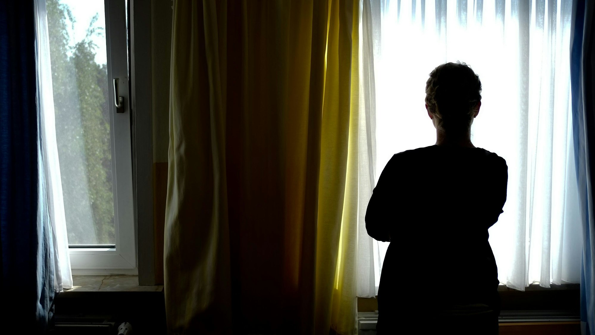 Eine Person steht vor einem Fenster und ist im Gegenlicht nur als dunkle Silhouette erkennbar.