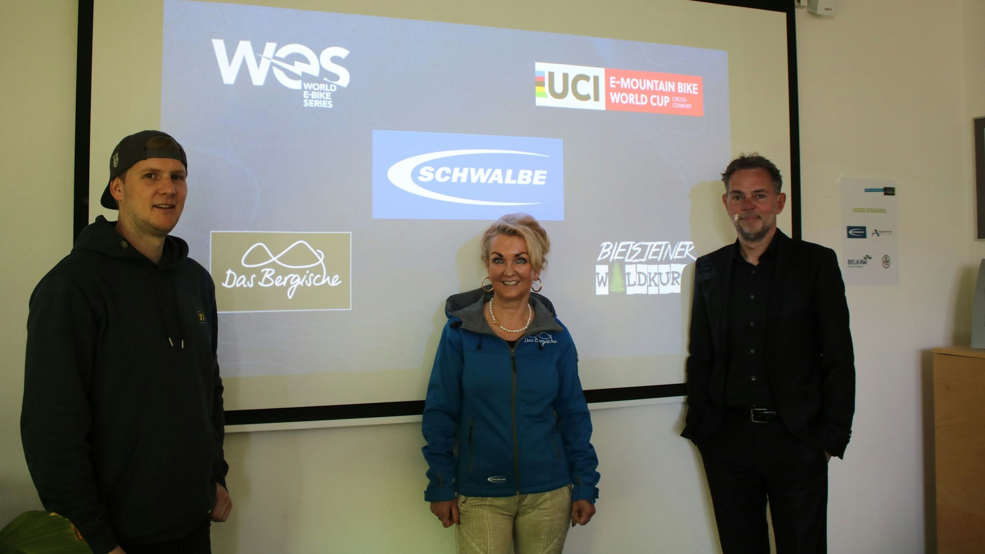 Zwei Männer und eine Frau stehen vor einer Leinwand mit mehreren Firmenlogos im Hintergrund.