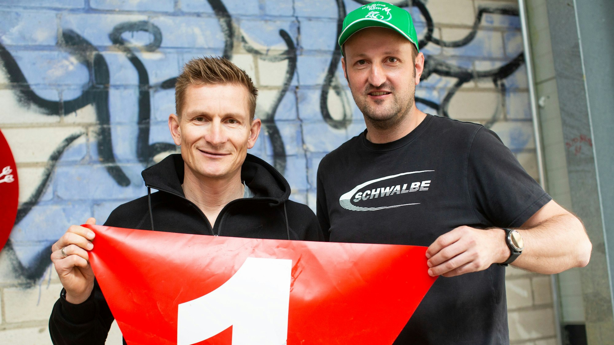 André Greipel (l.) als Sportlicher Leiter zeigt einen roten Wimpel in die Kamera.