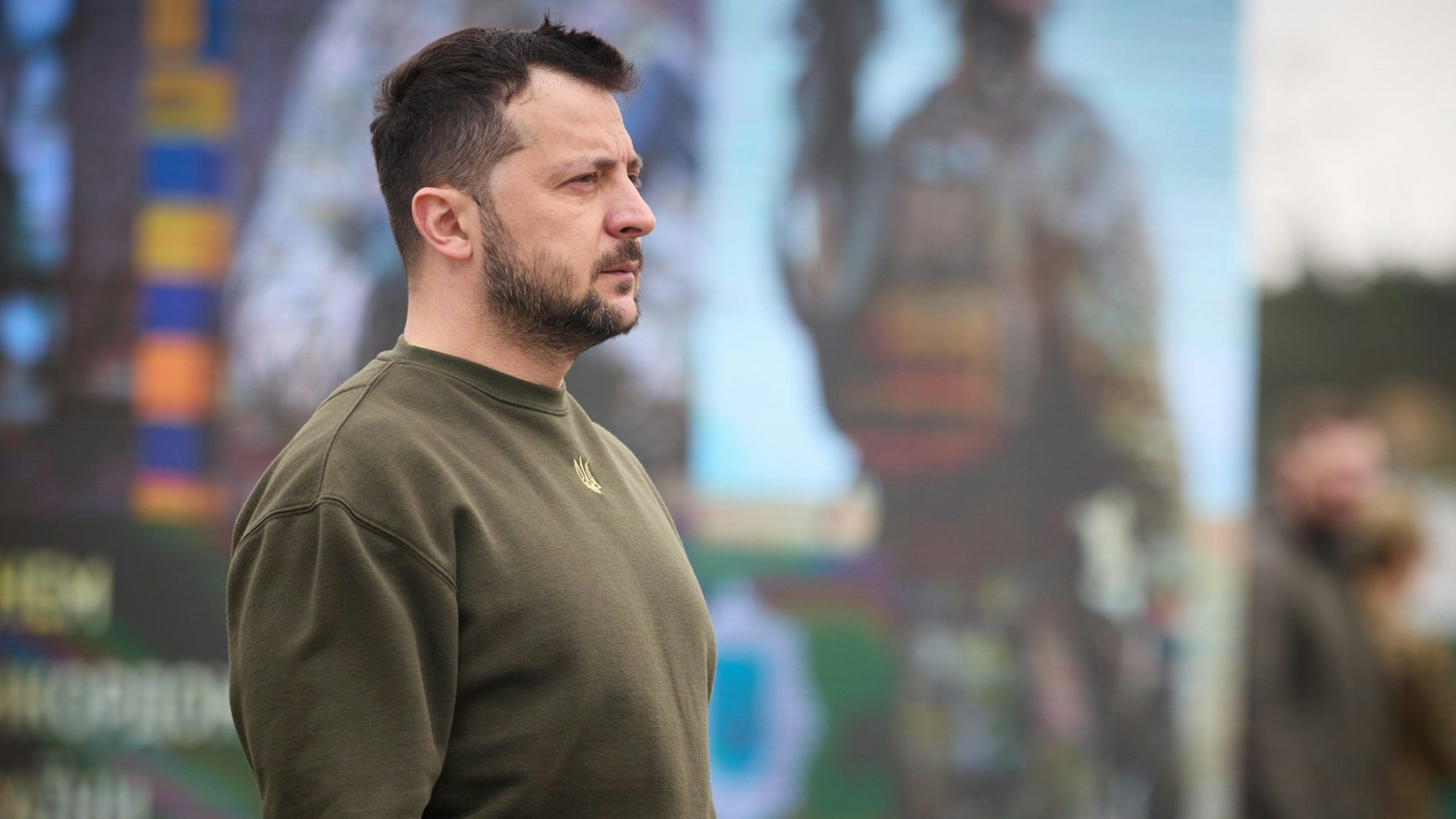 Der ukrainische Präsident Wolodymyr Selenskyj im Seitenprofil, er steht vor einem Plakat auf dem ein ukrainischer Soldat zu sehen ist.