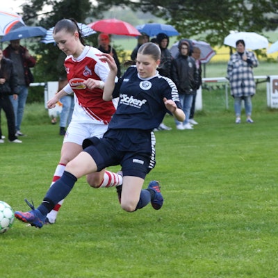 Eine Kommerner Spielerin setzt zu einer fairen Grätsche gegen eine Kölnerin an. Im Hintergrund stehen die Zuschauer mit Schirmen.