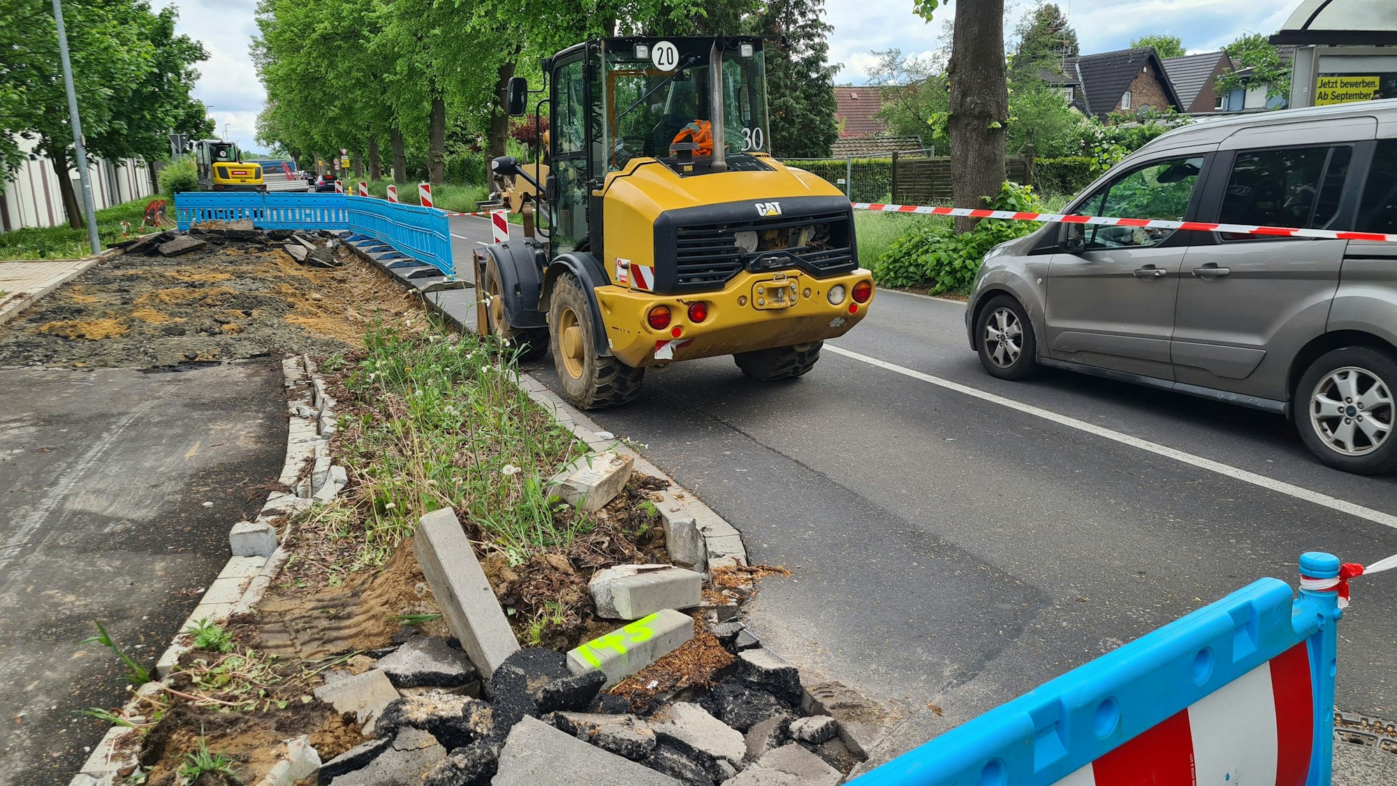Die Bushaltestelle an der Dansweilerstraße in Glessen wird dargestellt. Ein Baufahrzeug und ein Auto sind vordergründig an einer abgesperrten Baustelle zu sehen.
