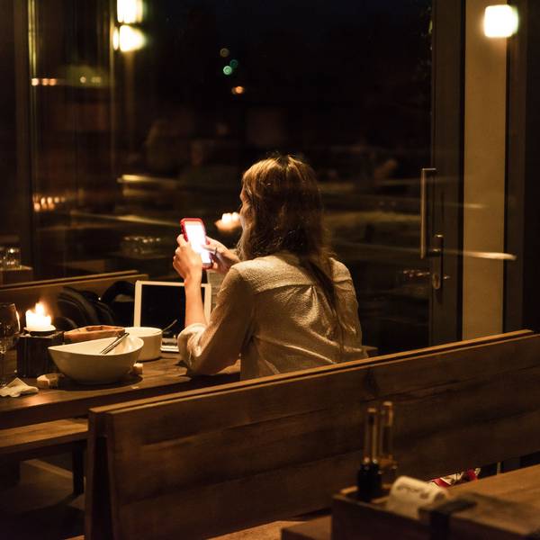 Eine Frau sitzt in einem relativ dunklen und weitestgehend leeren Restaurant an einem Tisch mit leerem Teller und schaut auf ihr Smartphone.