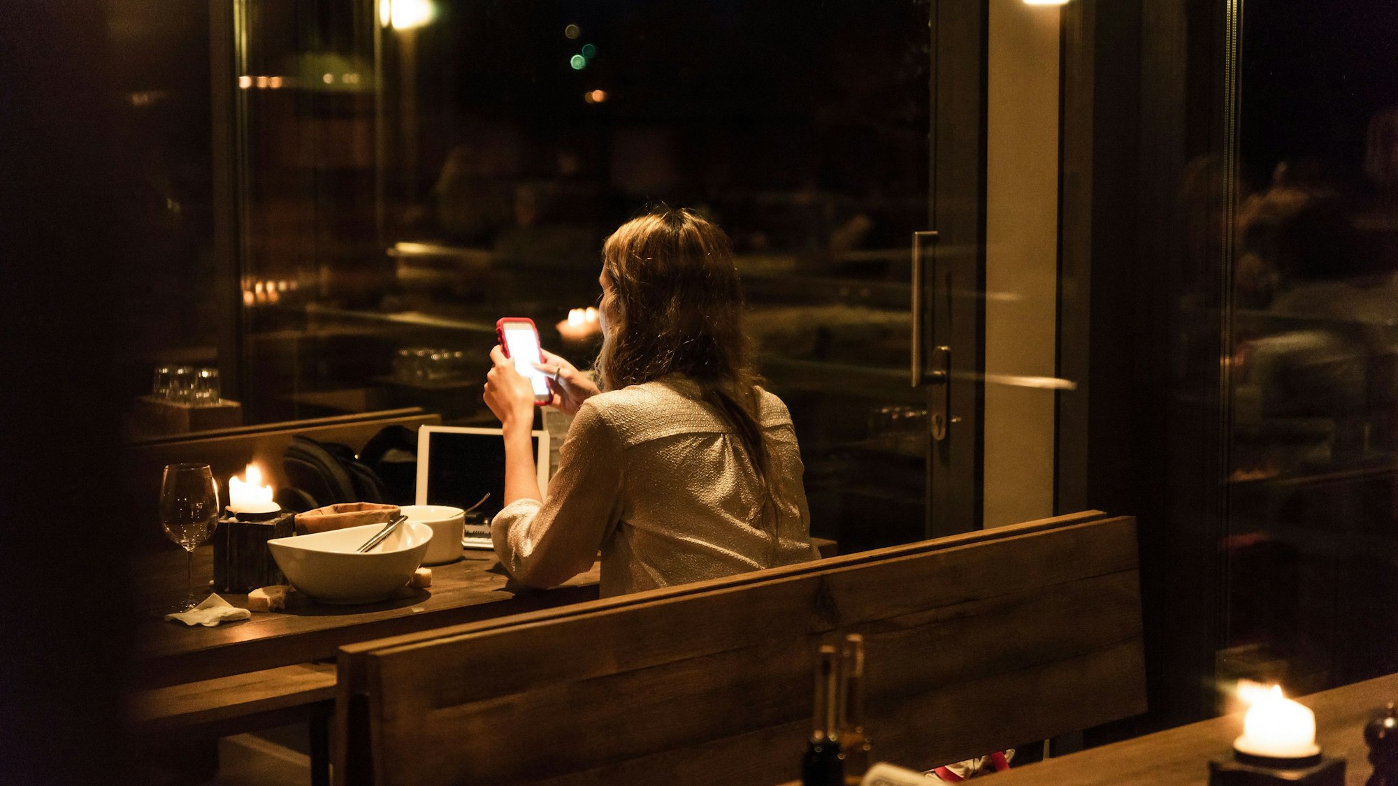 Eine Frau sitzt in einem relativ dunklen und weitestgehend leeren Restaurant an einem Tisch mit leerem Teller und schaut auf ihr Smartphone.
