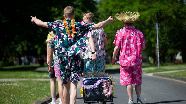 Junge Männer in Outfits mit floralen Mustern gekleidet, ziehen einen geschmückten Bollerwagen mit Getränken am Aachener Weiher hinter sich her.