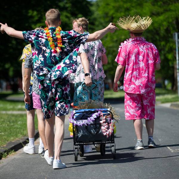 Junge Männer in Outfits mit floralen Mustern gekleidet, ziehen einen geschmückten Bollerwagen mit Getränken am Aachener Weiher hinter sich her.