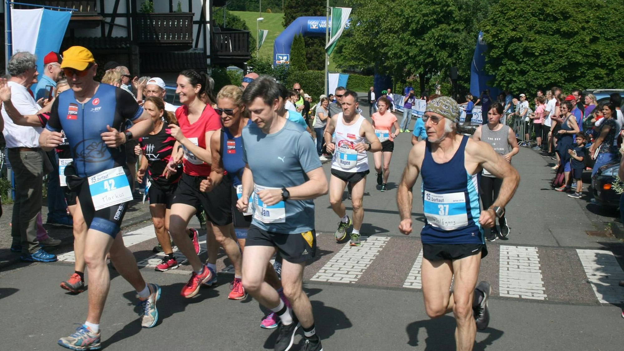 Der Biesfelder Dorflauf 2019. Mehrere Läufer und Läuferinnen in Sportklamotten sind zu sehen. Publikum feuert die Athleten und Athletinnen an.