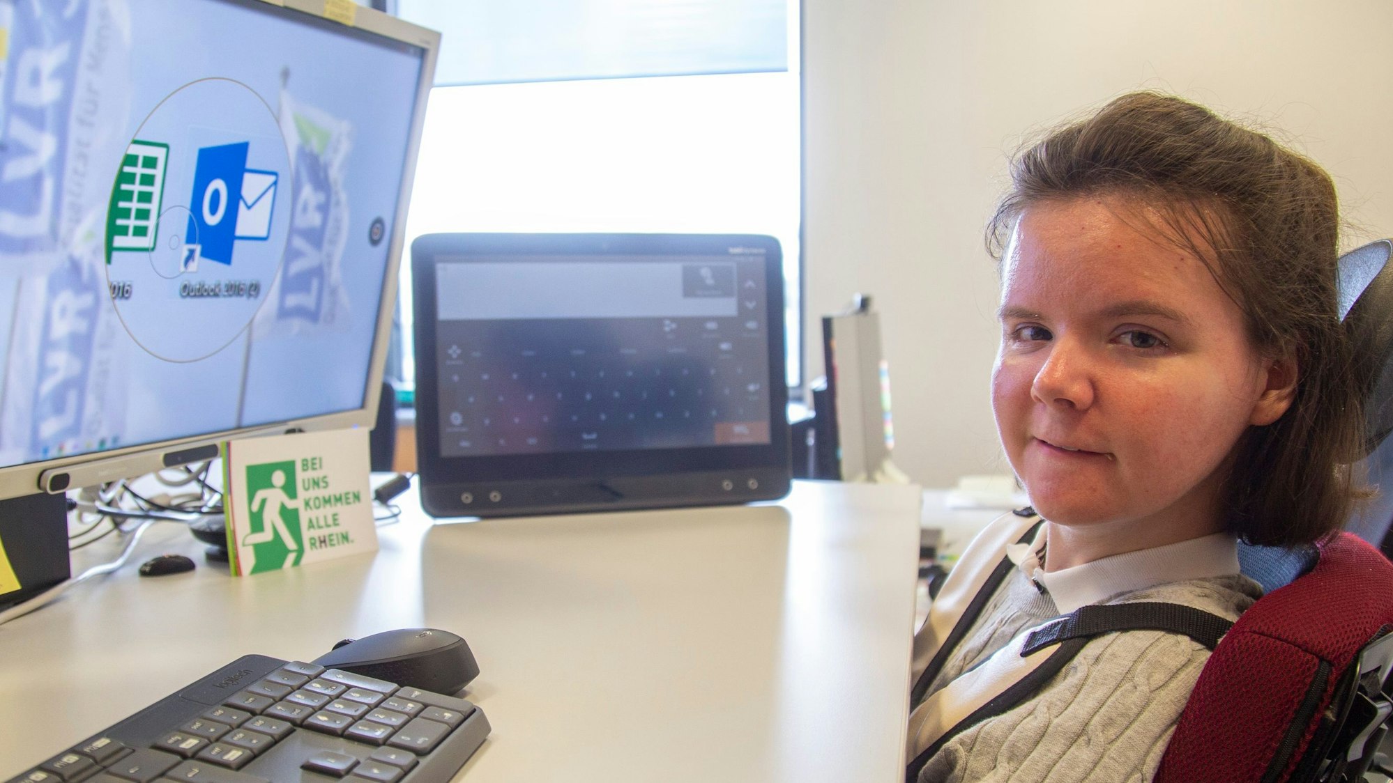 Julia Holler sitzt in einem Rollstuhl direkt vor ihrem Schreibtisch. Auf dem Schreibtisch stehen drei Bildschirme.