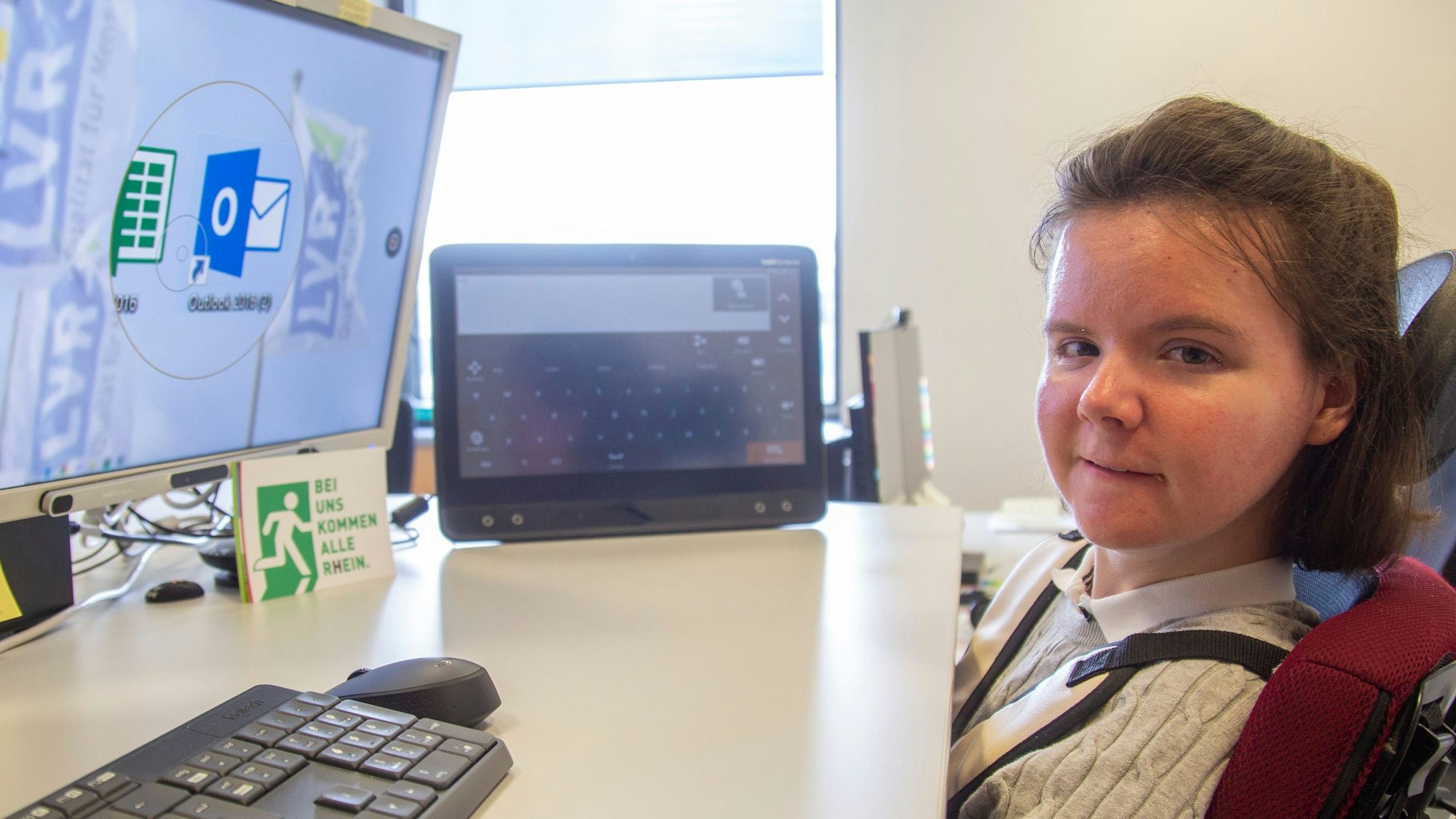 Julia Holler sitzt in einem Rollstuhl direkt vor ihrem Schreibtisch. Auf dem Schreibtisch stehen drei Bildschirme.