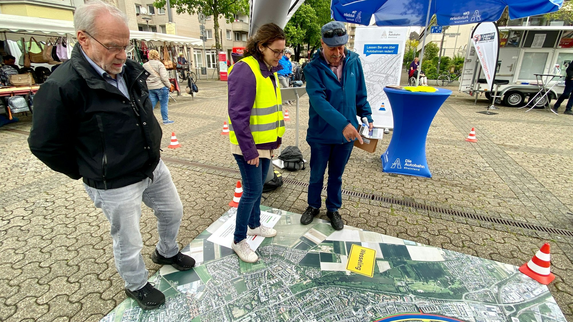 Mitarbeiter der Autobahn GmbH stehen auf einem großen Plakat, welches die A555 und Umgebung zeigt.