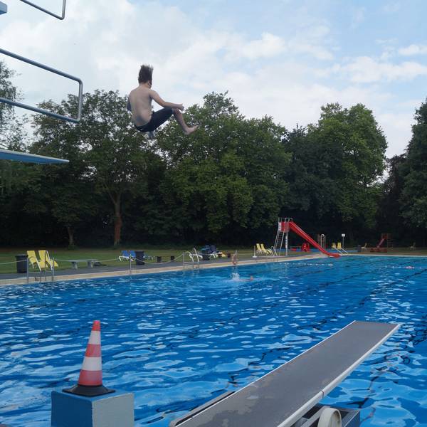 Ein Junge springt von einem drei-Meter-Brett in ein großes Außenschwimmbecken. Im Hintergrund sind eine Rutsche und Bäume zu sehen.