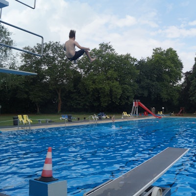 Ein Junge springt von einem drei-Meter-Brett in ein großes Außenschwimmbecken. Im Hintergrund sind eine Rutsche und Bäume zu sehen.