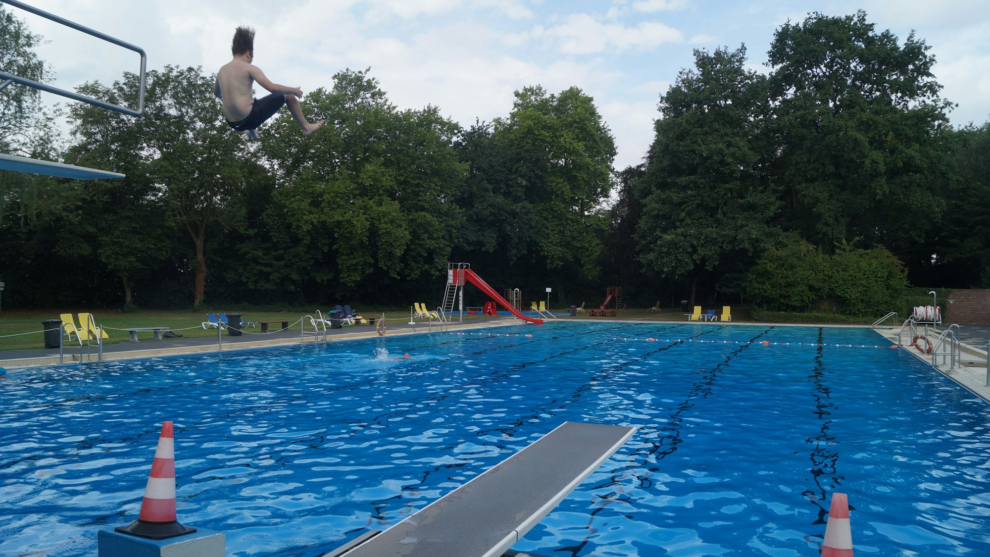 Ein Junge springt von einem drei-Meter-Brett in ein großes Außenschwimmbecken. Im Hintergrund sind eine Rutsche und Bäume zu sehen.
