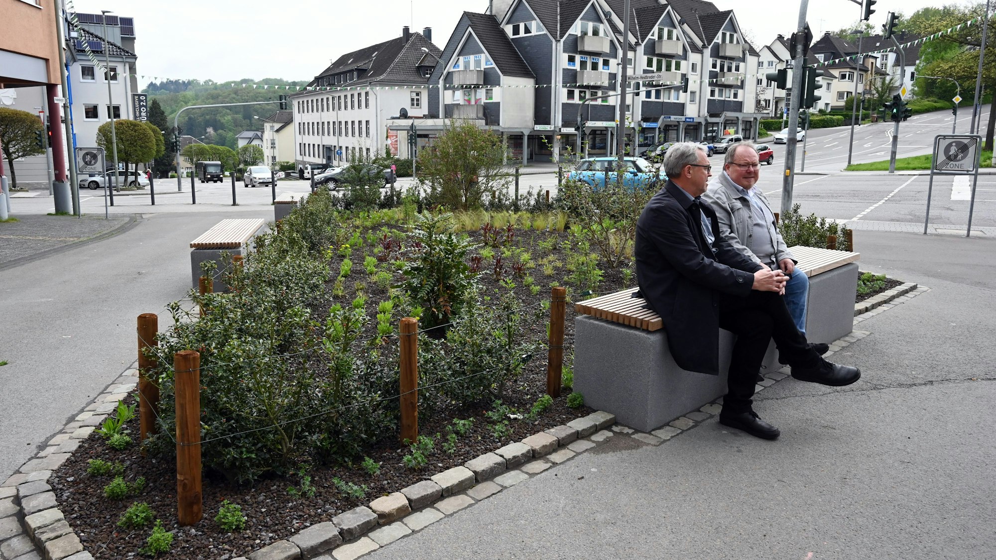 Zwei Männer sitzen auf einer Bank an einer Stadt-Kreuzung. Hinter ihnen ist ein Beet zu sehen.