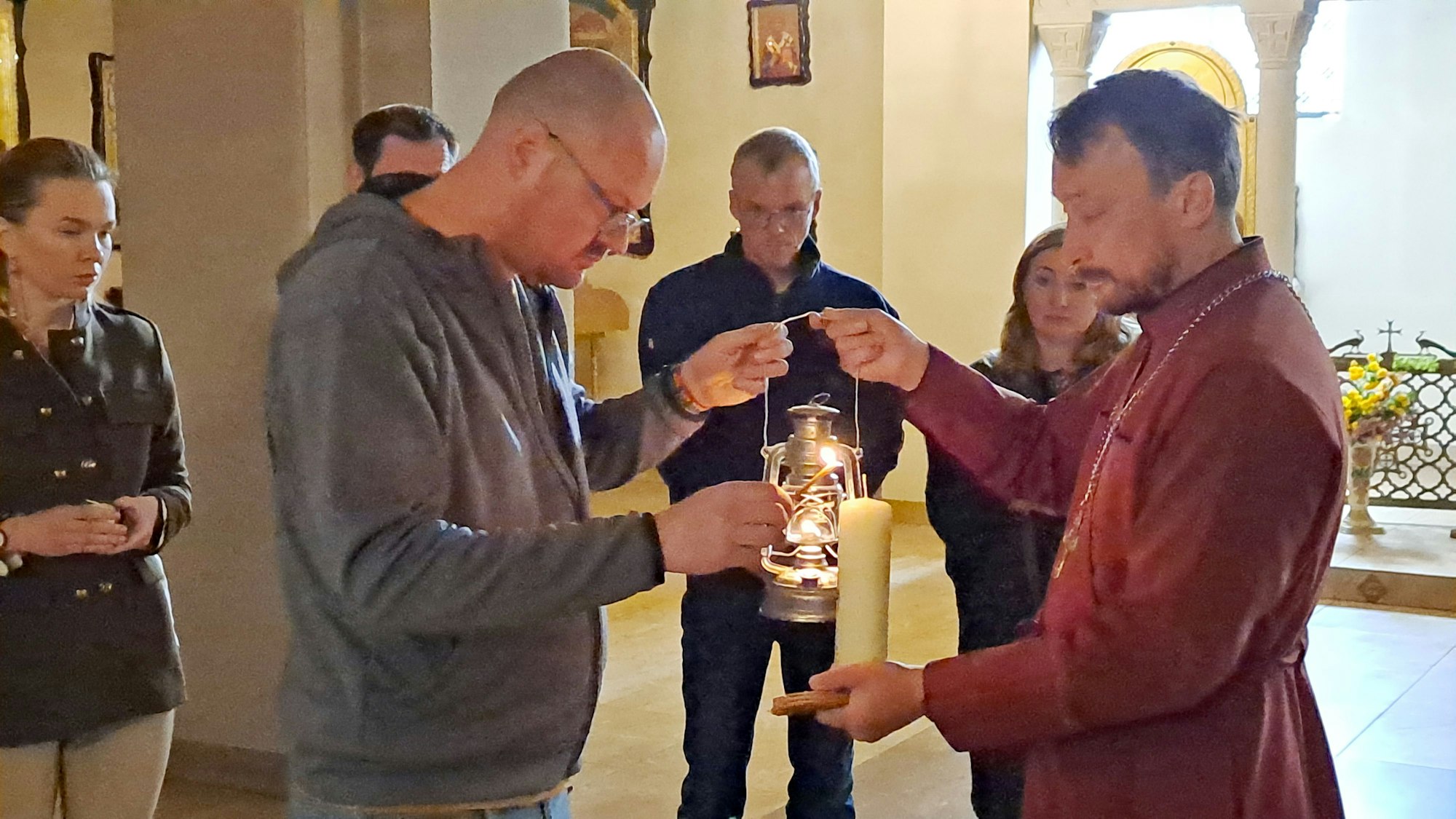 Ein Mann zündet an einer Laterne einen Docht an, mit dem er die Kerze eine Gesitlichen entzündet.