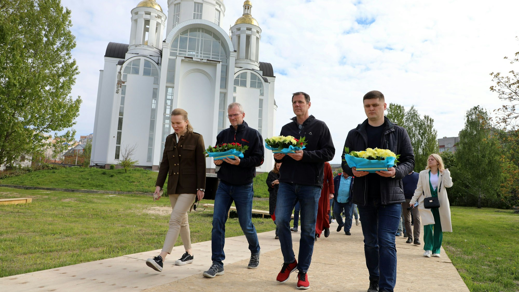 Menschen mit Blumen in den Händen gehen einen Holfweg hinter einer orthodoxen Kirche, der Andreas-Kirche, in Butscha antlang zu einer Gedenkstätte.
