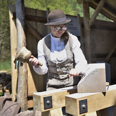 Steinmetzin Ulrike Glaubitz steht vor der Steinmetzhütte im Freilichtmuseum Kommern. Sie trägt traditionelle, beige Handwerks-Kluft und bearbeitet einen Stein mit Hammer und Meißel.