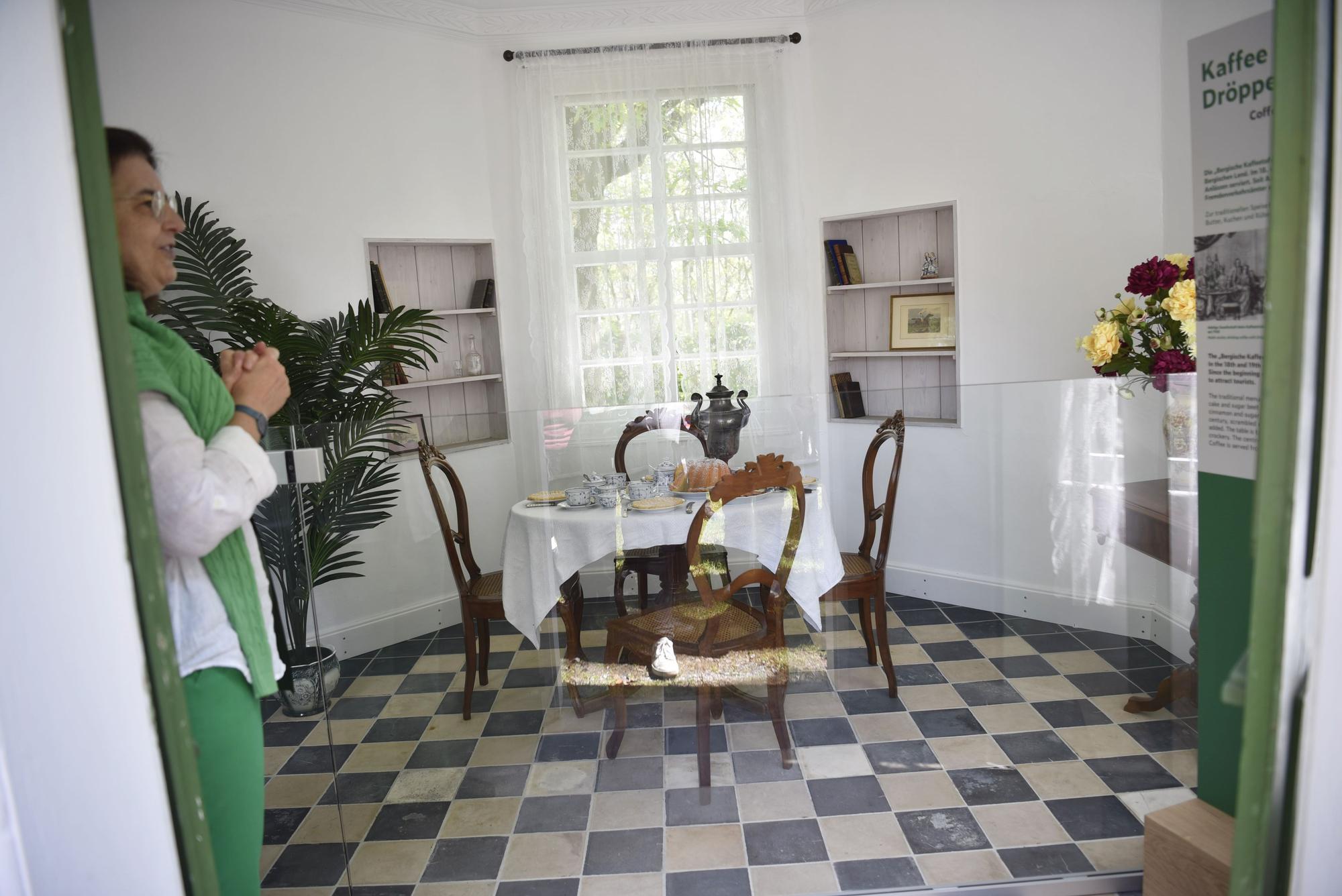 In einem fünfeckigen Raum stehen vor einem Fenster ein Tisch und vier Stühle. Auf dem Tisch liegt eine weiße Tischdecke. Der Tisch ist mit blau-weißem Geschirr gedeckt. Es sind ein Kuchen und eine Dröppelminna (eine alte Kaffeekanne) zu sehen. Am Rand stehen Blumen.