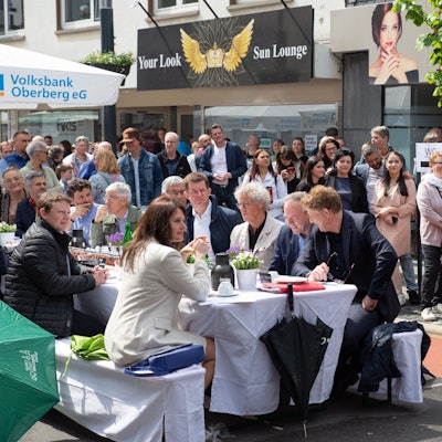 Zahlreiche Menschen säumen eine Straße in einem Stadtzentrum. In der Mitte steht eine lange weiße Tafel, an der ebenfalls viele Menschen sitzen.