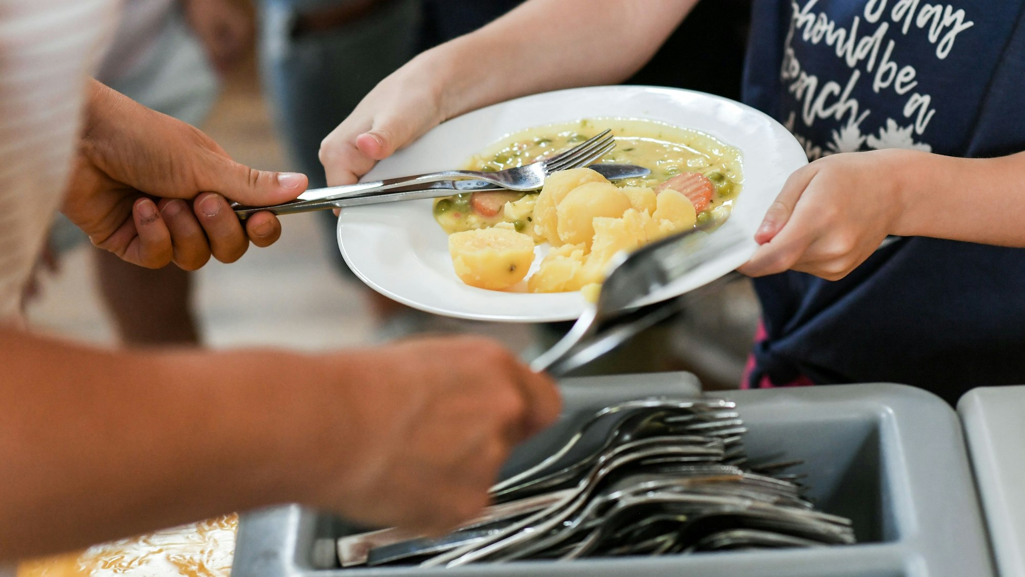 Das Foto zeigt Schüler beim Mittagessen in der Mensa in einer Grundschule. Im Vordergrund ist ein Besteckkasten zu sehen.